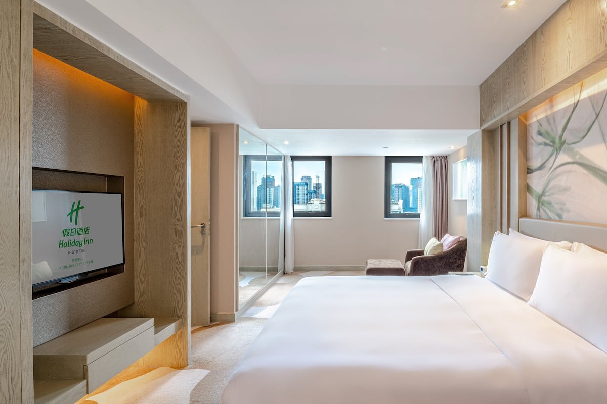 Ein Hotelzimmer mit einem großen Bett, einem Fernseher links und Blick auf die Stadt durch die Fenster.