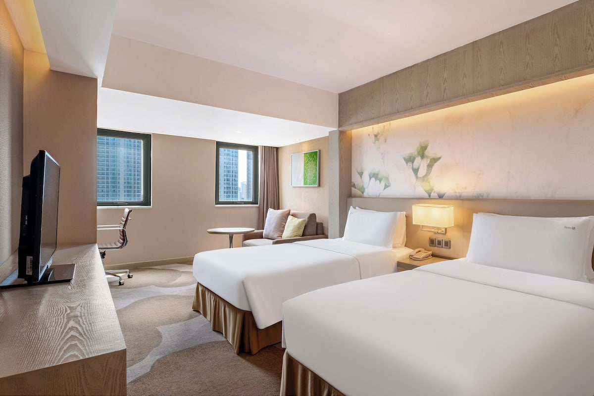 Ein Hotelzimmer mit zwei Betten, einem Schreibtisch, einem Fernseher und Blick auf ein Gebäude.