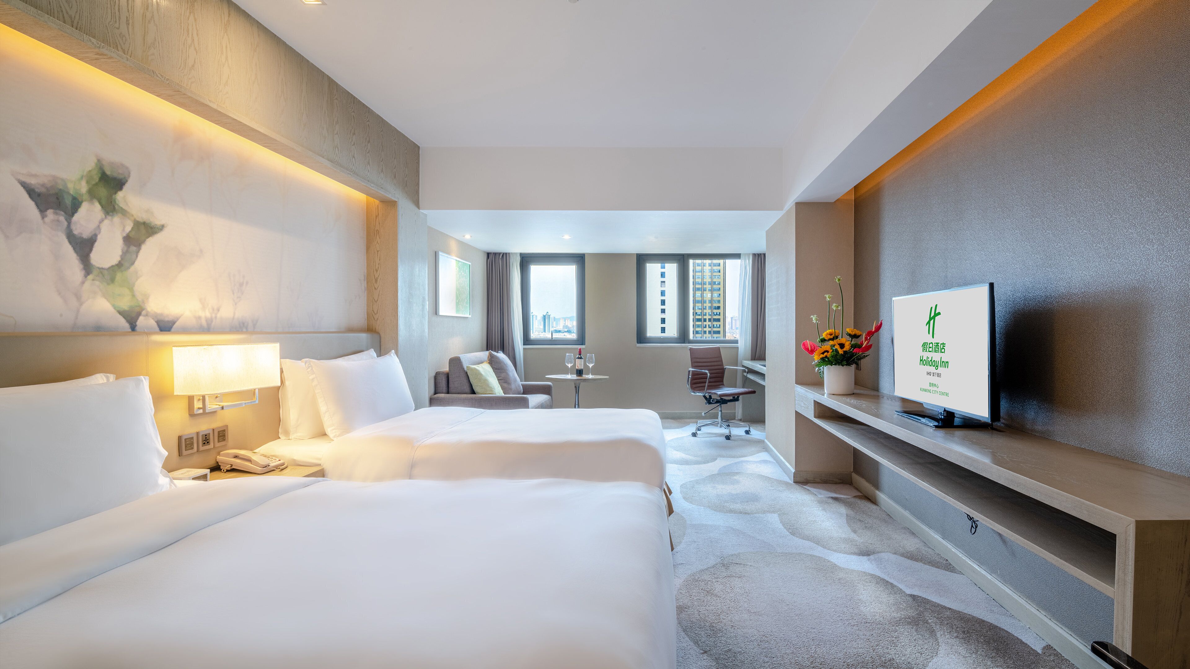 A hotel room with two beds, a desk, a chair, a TV, and a view of the city.
