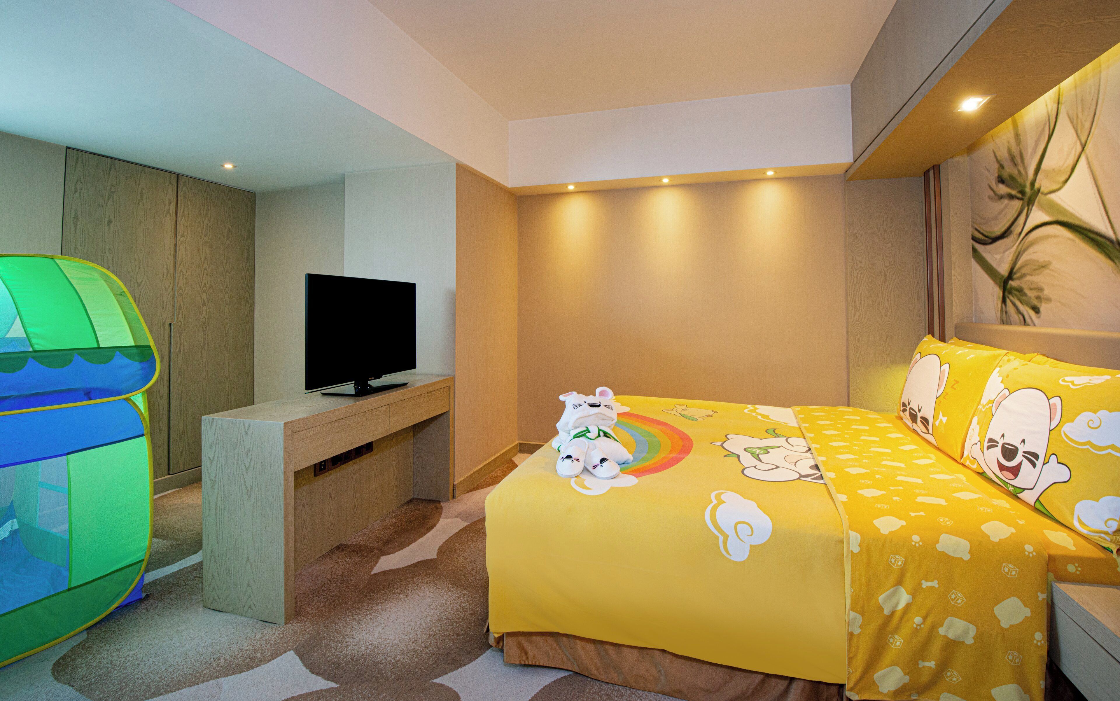 A bedroom with a bed, a television on a stand, a green play tent, and a wall with a plant.