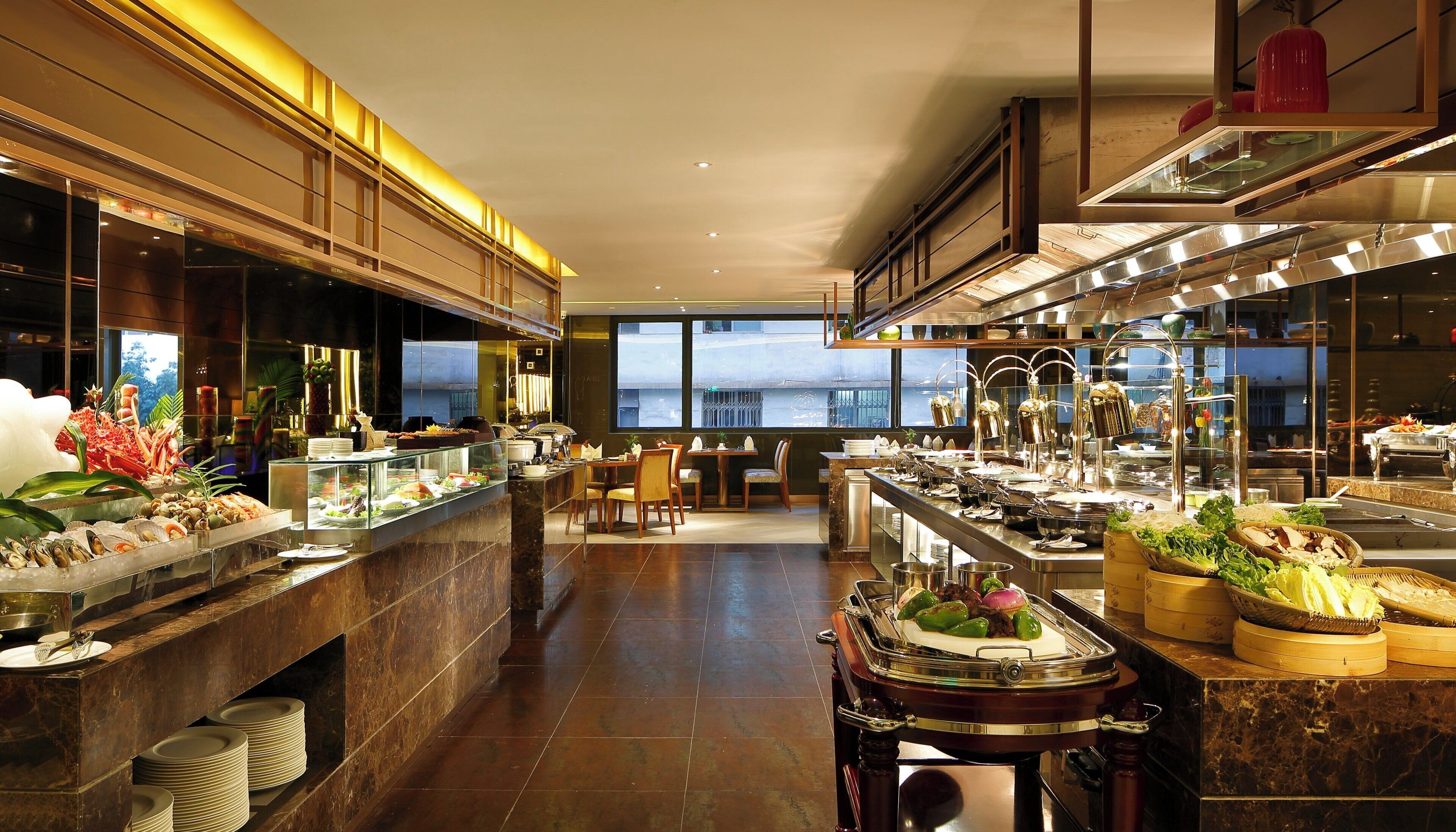 A modern hotel dining area with a buffet setup, a seating area, and a view of the cityscape through large windows.