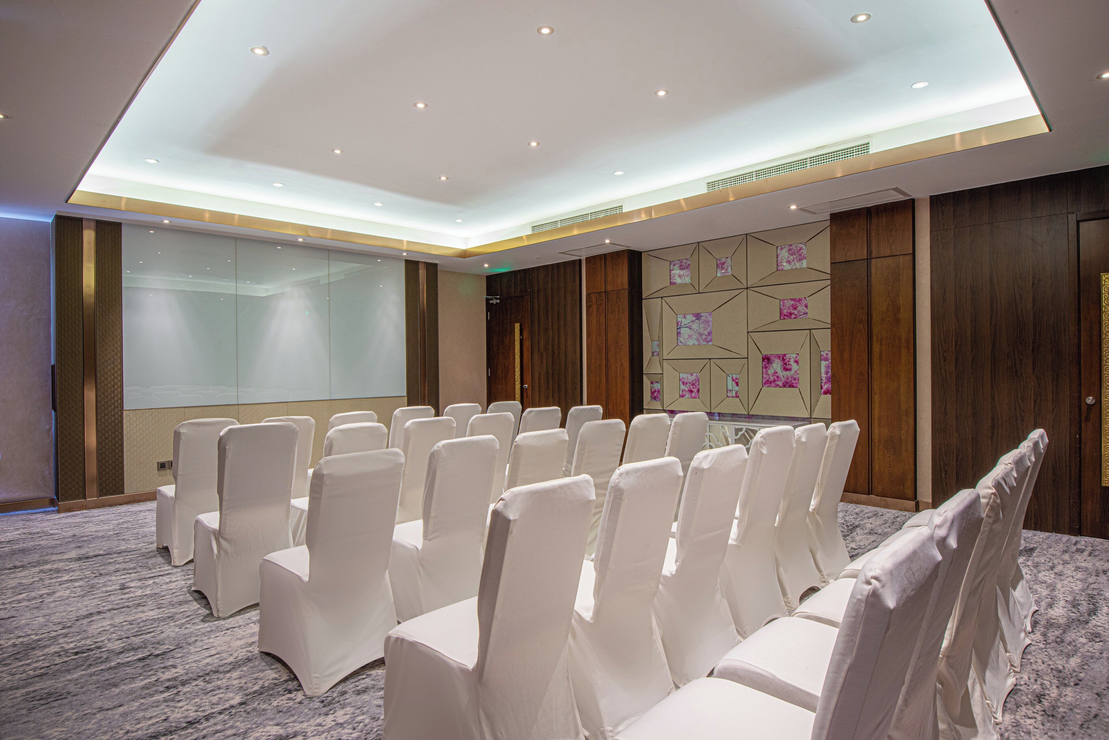 Ein Konferenzraum mit Reihen weißer Stühle, einer Projektionsleinwand und einer Wand mit geometrischen Mustern.