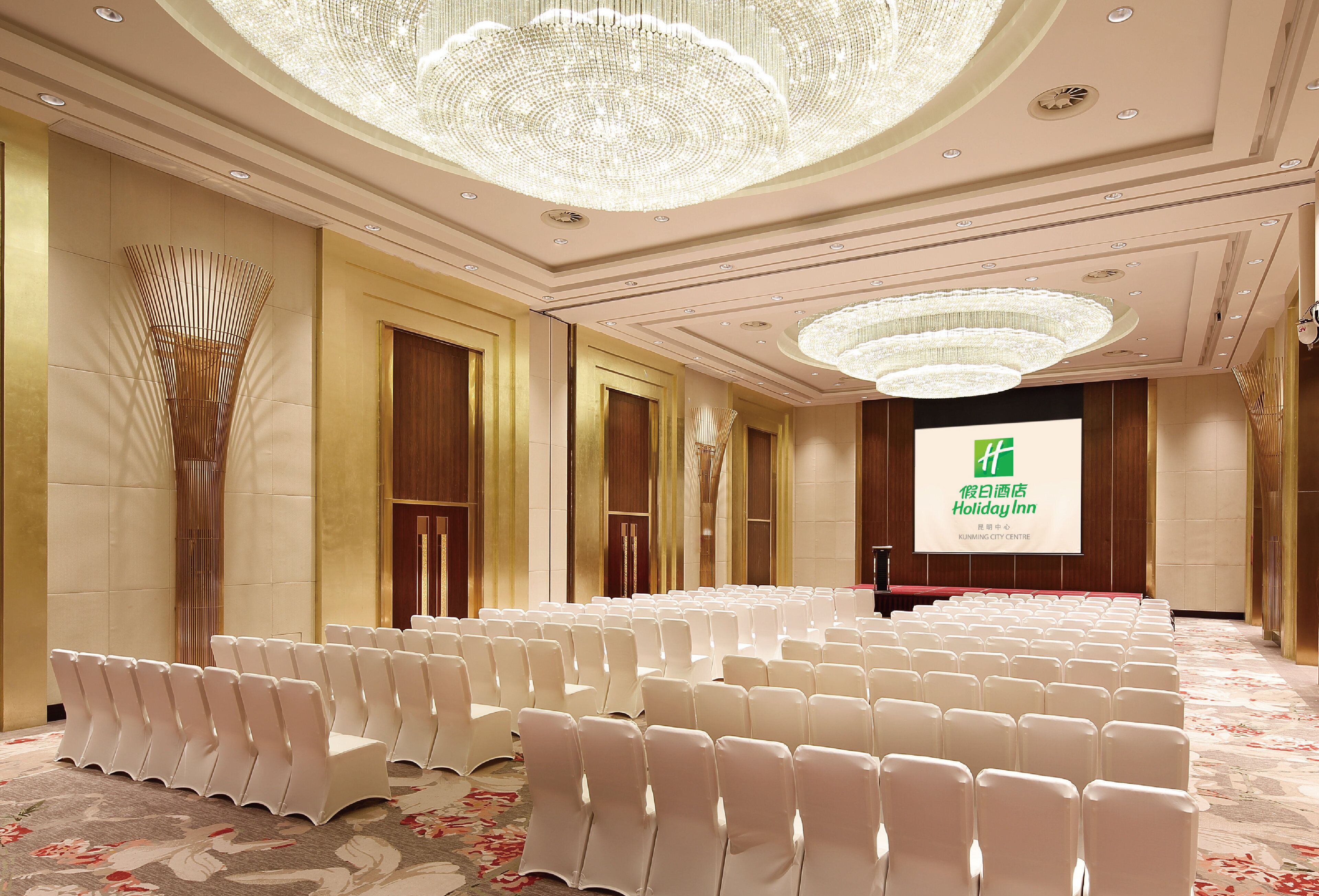 Un salón de conferencias con hileras de sillas blancas, una lámpara de araña grande y una pantalla con el logotipo de Holiday Inn.