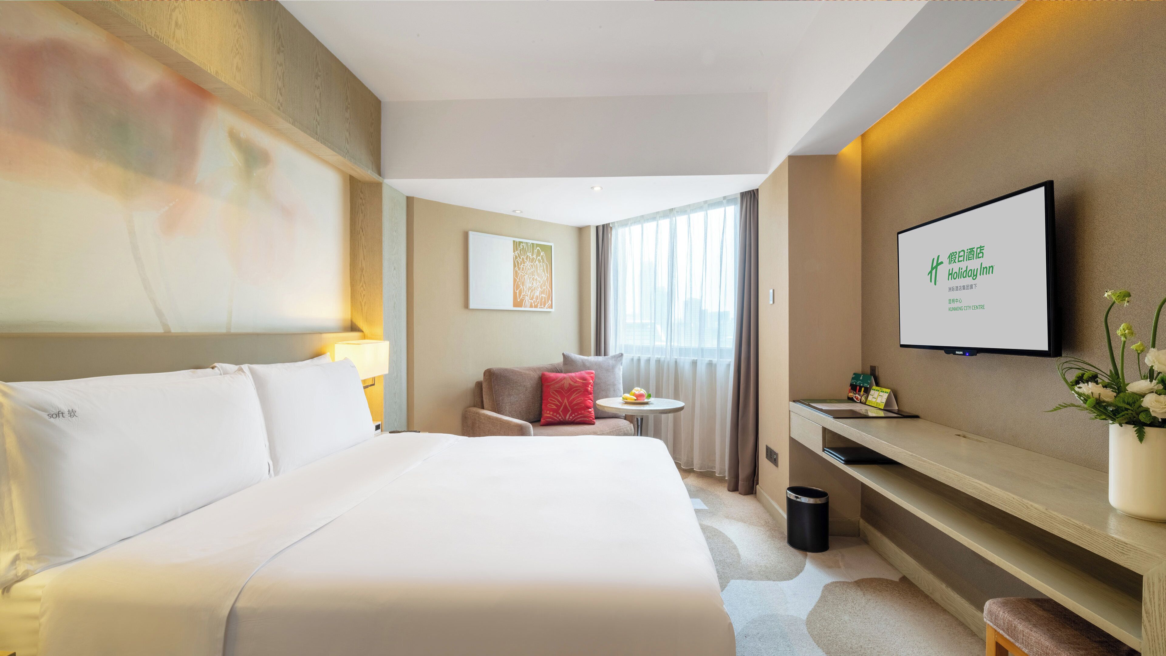 Ein Hotelzimmer mit einem großen Bett, einem Schreibtisch, einem Stuhl, einem kleinen Tisch mit Lampe, einem an der Wand befestigten Fernseher und einem Fenster mit Vorhängen.