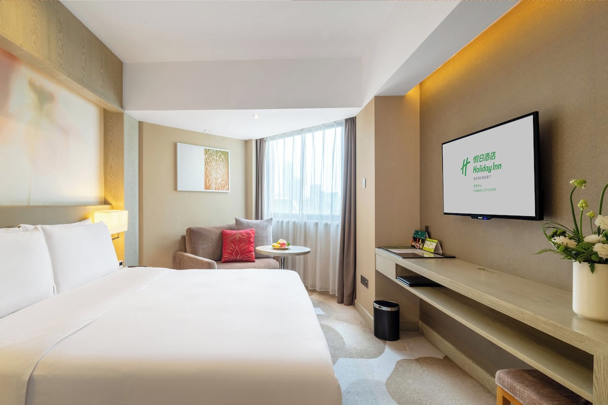Ein Hotelzimmer mit einem großen Bett, einem Schreibtisch, einem Stuhl, einem kleinen Tisch mit Lampe, einem an der Wand befestigten Fernseher und einem Fenster mit Vorhängen.