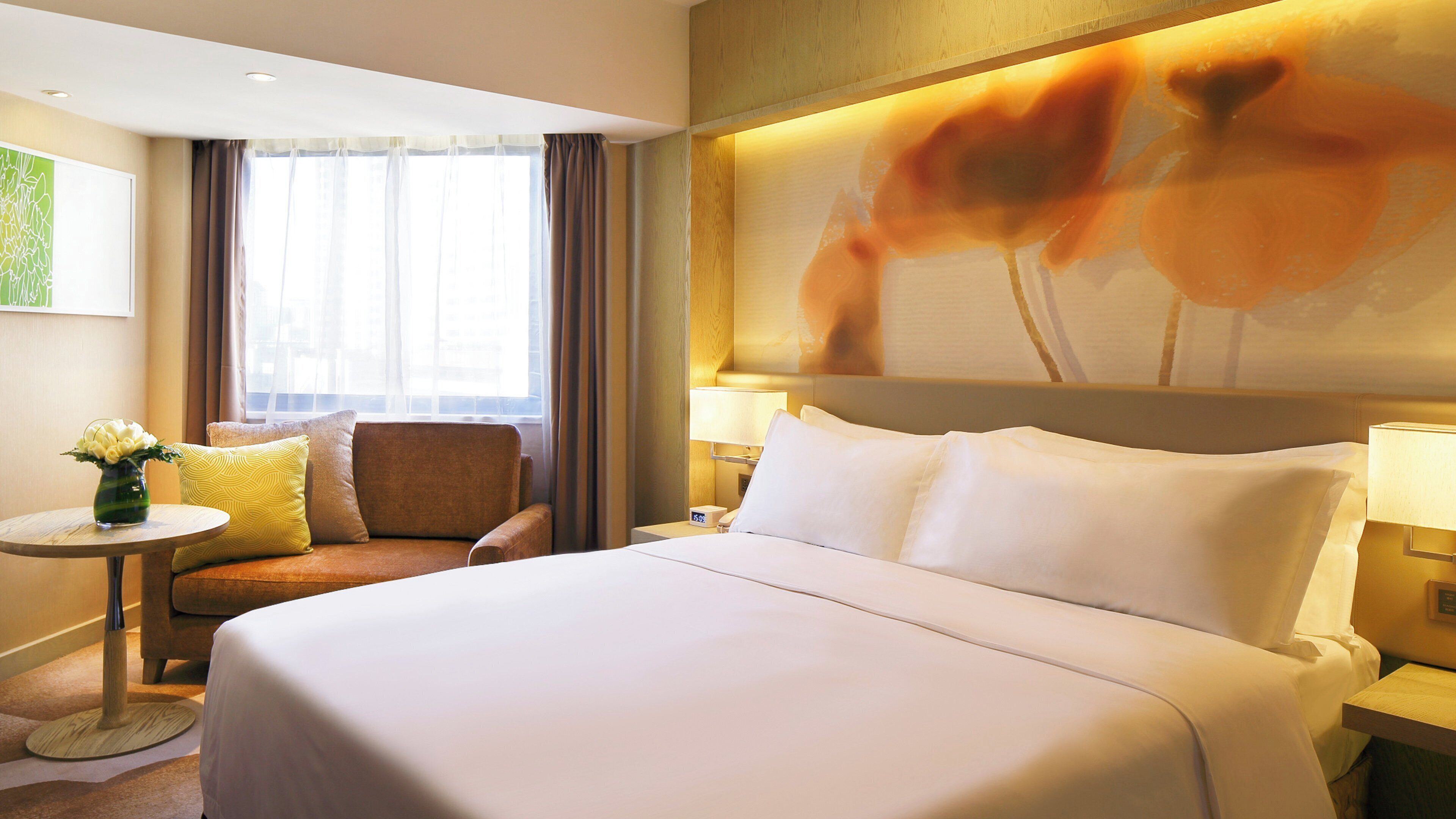 A hotel room with a bed, a chair, a small table with a vase, and a painting on the wall.