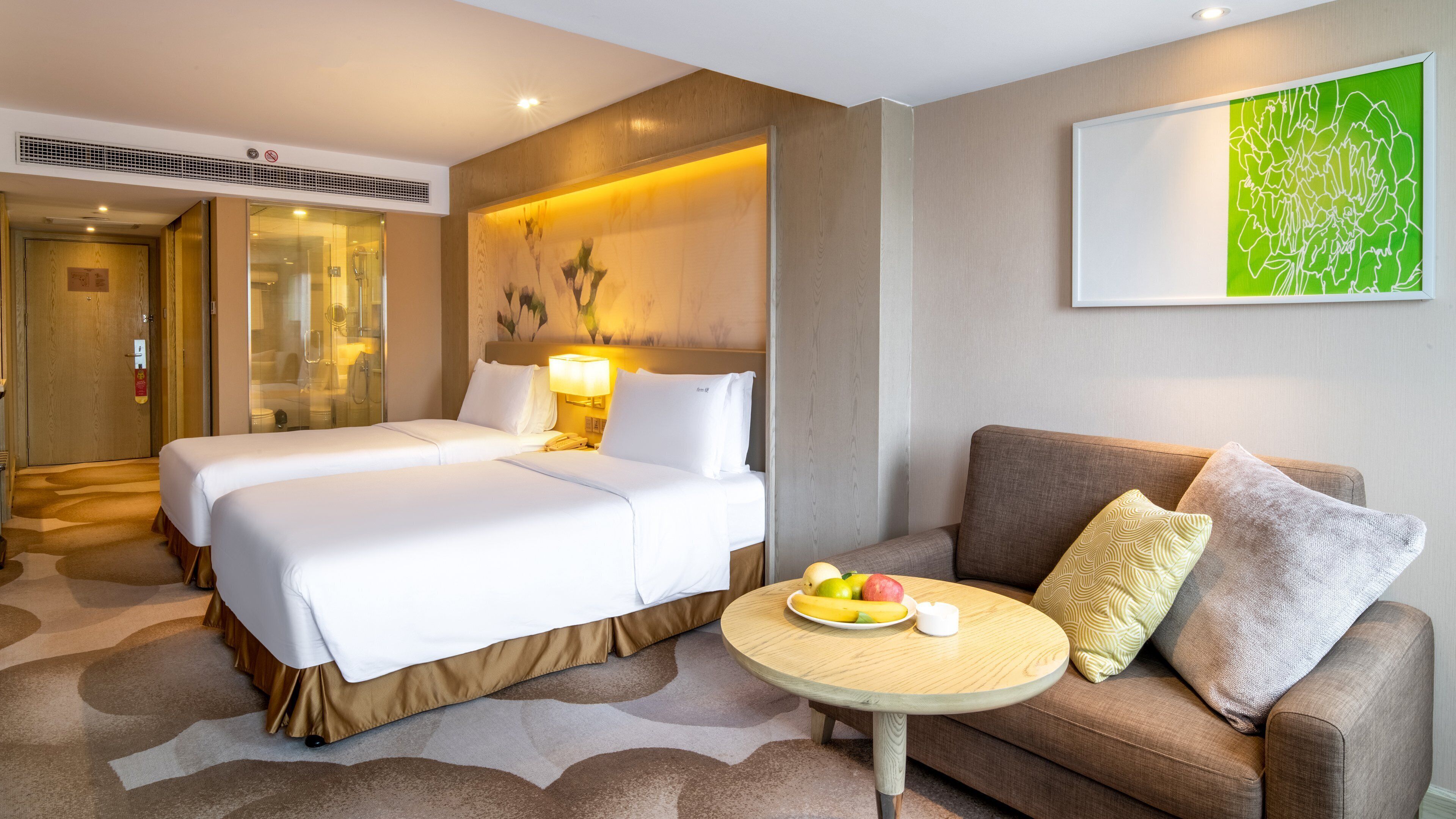 Hotelkamer met een groot bed, een bank, een klein rond tafeltje met fruit en een abstract schilderij in groene tinten aan de muur.