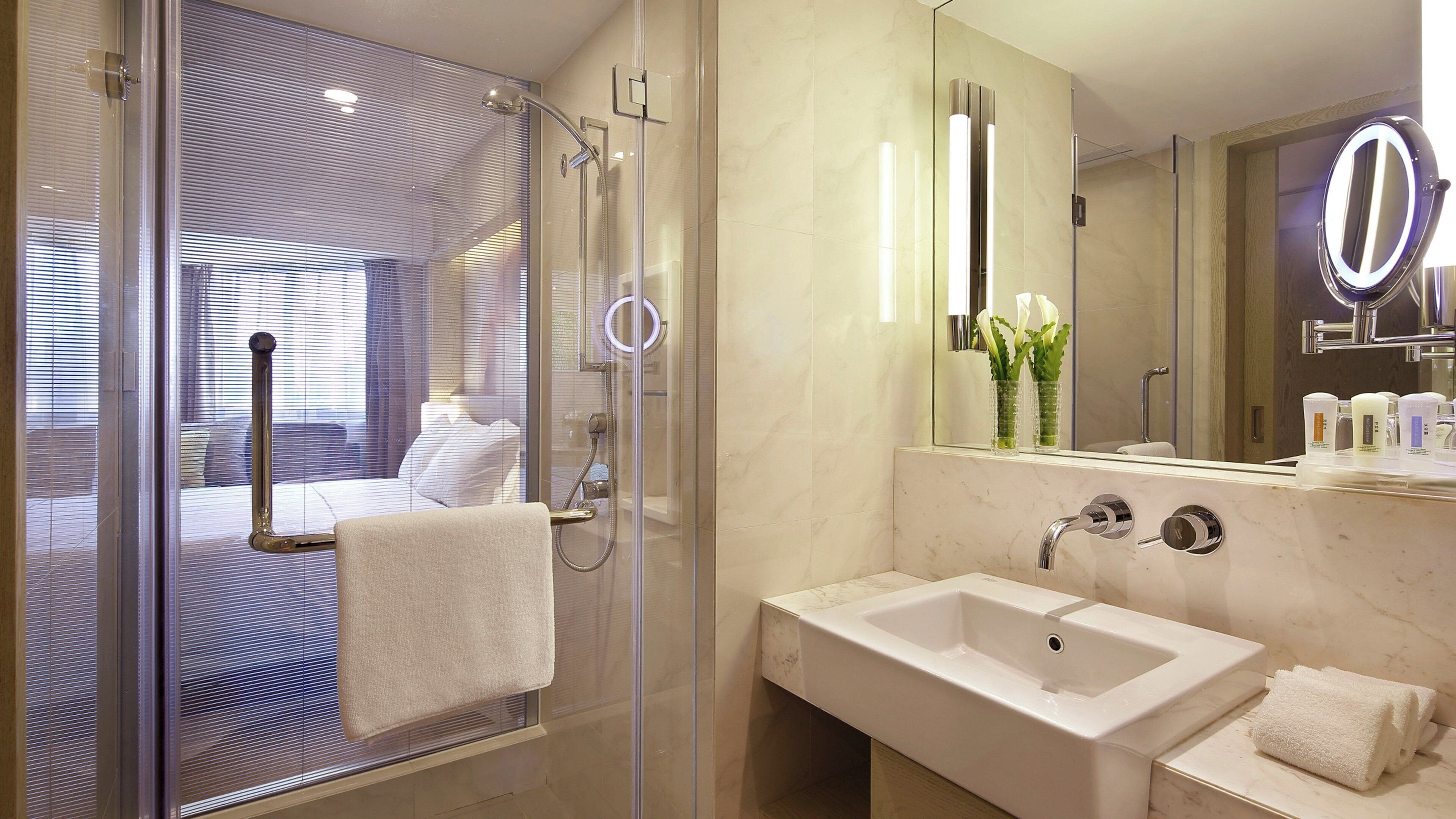 Un baño moderno con ducha envuelta en cristal, un espejo grande con marco iluminado y un lavamanos blanco.