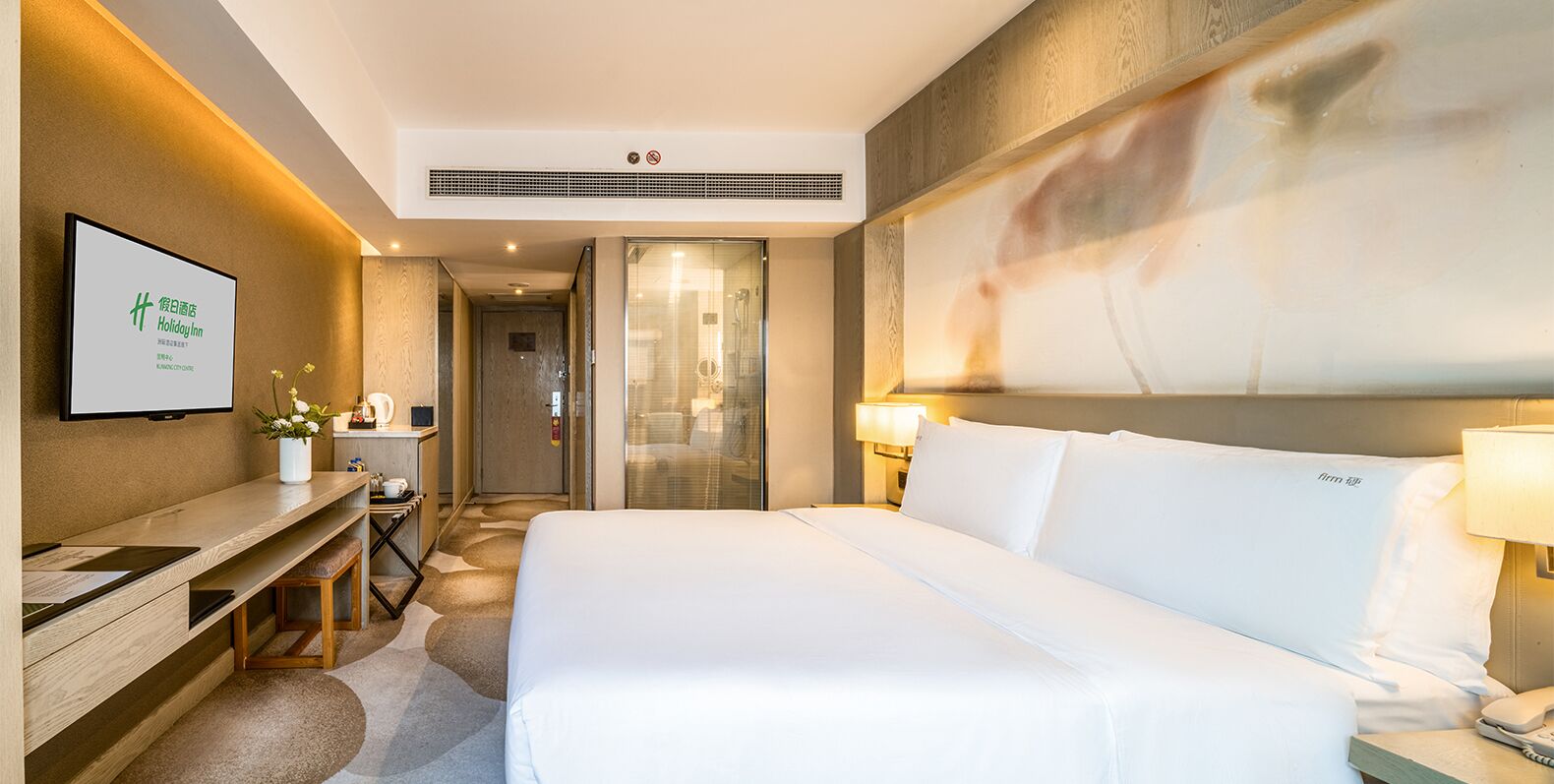 A hotel room with a bed, bedside table, and a desk with a chair. There is a shower area visible through a glass door.