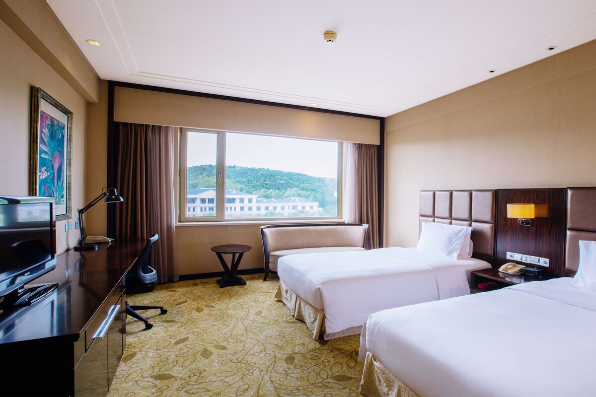 Habitación de hotel con dos camas, un escritorio con televisor, una lámpara y vista a un edificio desde la ventana.