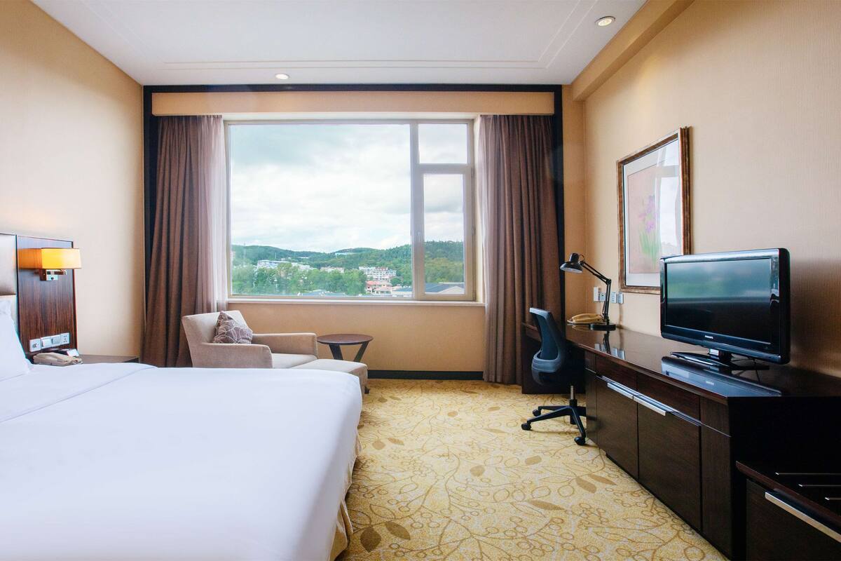 A hotel room with a large bed, a desk with a television, a chair, a small table, and a window with a view of the city.