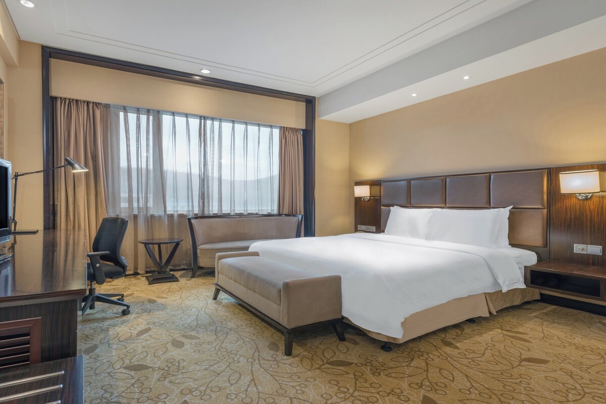 Una habitación de hotel con una cama grande, un escritorio con televisor de pantalla plana, una silla, una mesita y una ventana con cortinas.