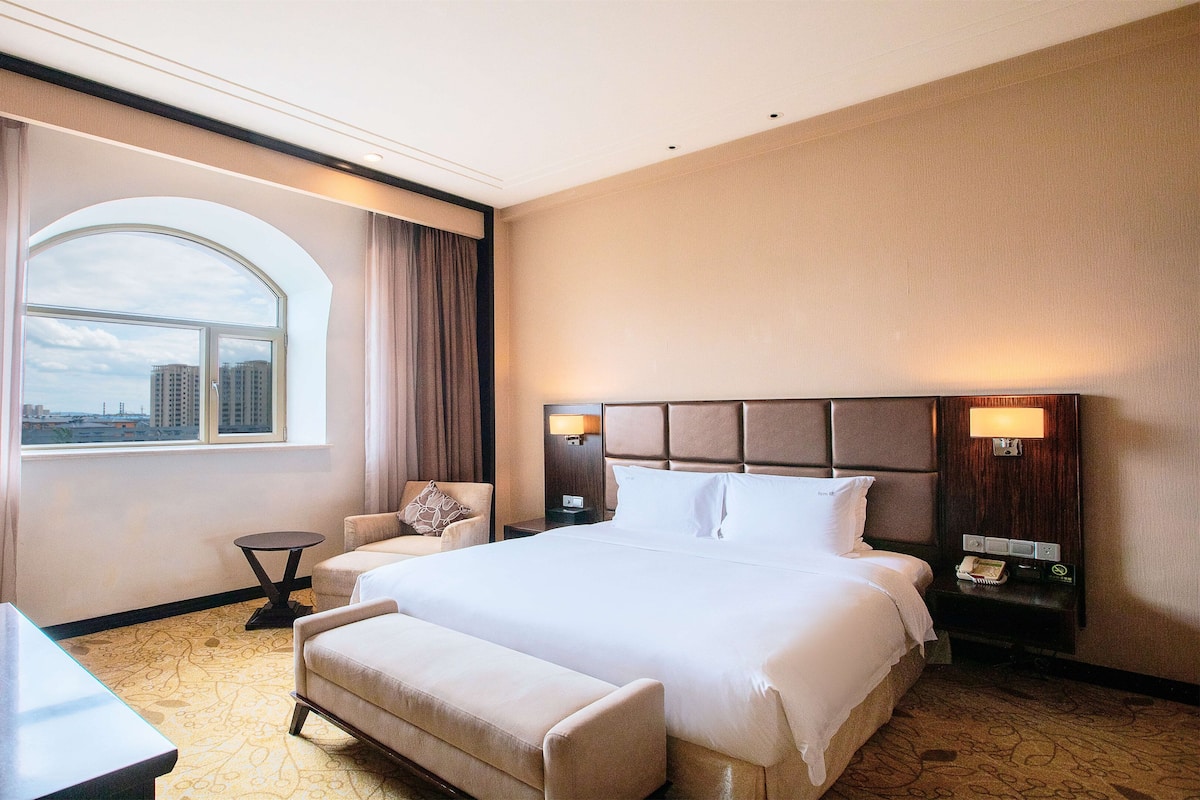 A hotel room with a large bed, a desk, a chair, and a view of the city through a large window.