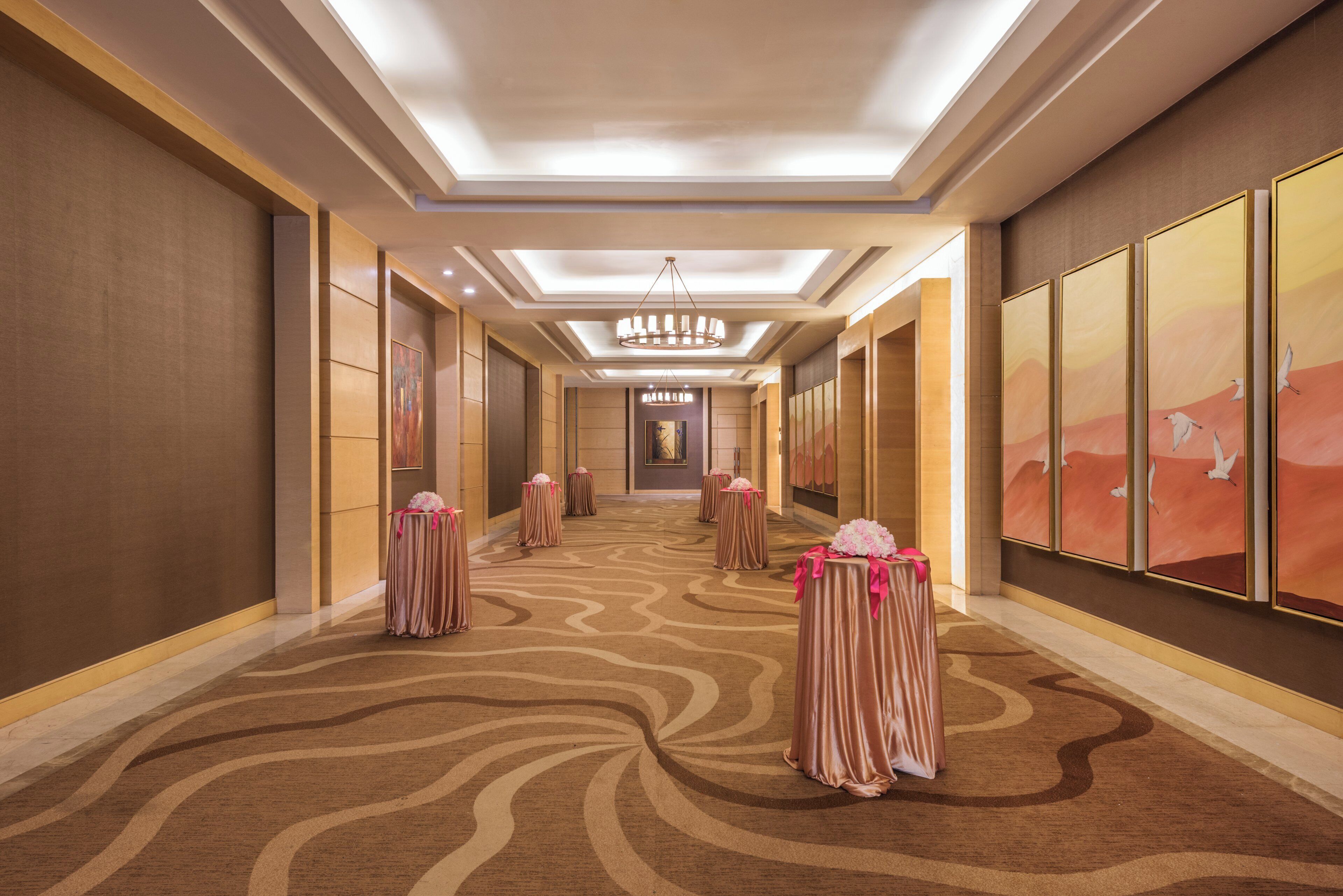 A hotel corridor with a patterned carpet, a chandelier, and framed artwork on the walls.