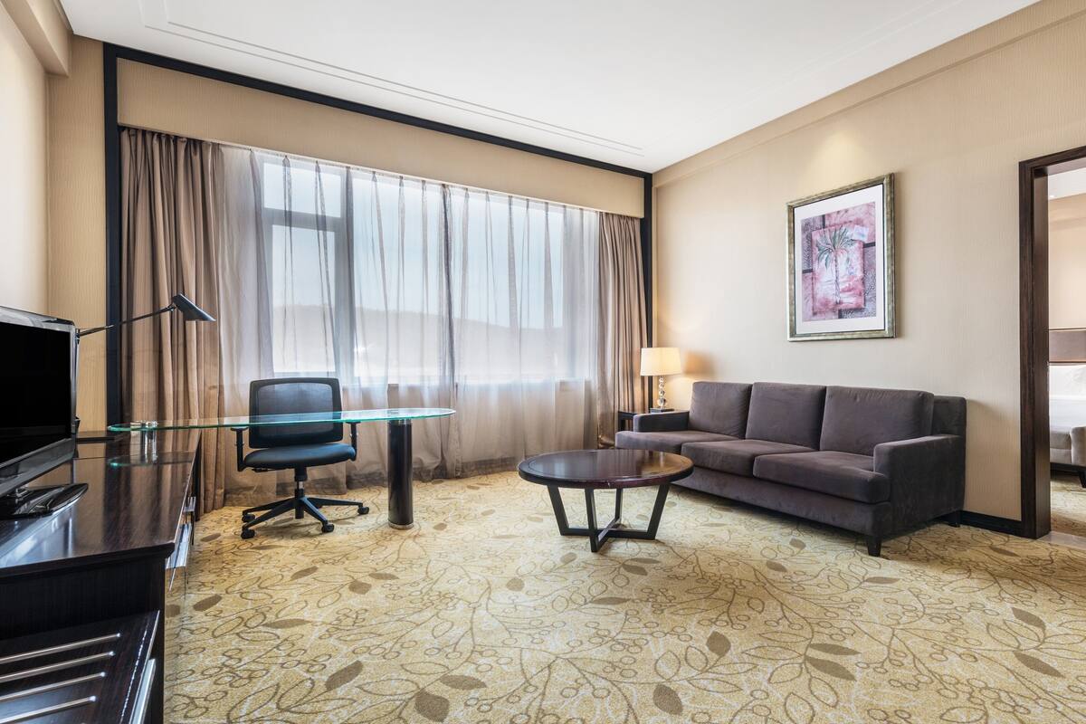 A hotel room with a glass desk, a sofa, a coffee table, a TV, and a bed.