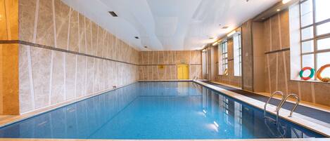 Una piscina interior con agua azul cristalina, paredes y piso de azulejos. Hay dos puertas, una amarilla y otra blanca, y una ventana a la derecha.