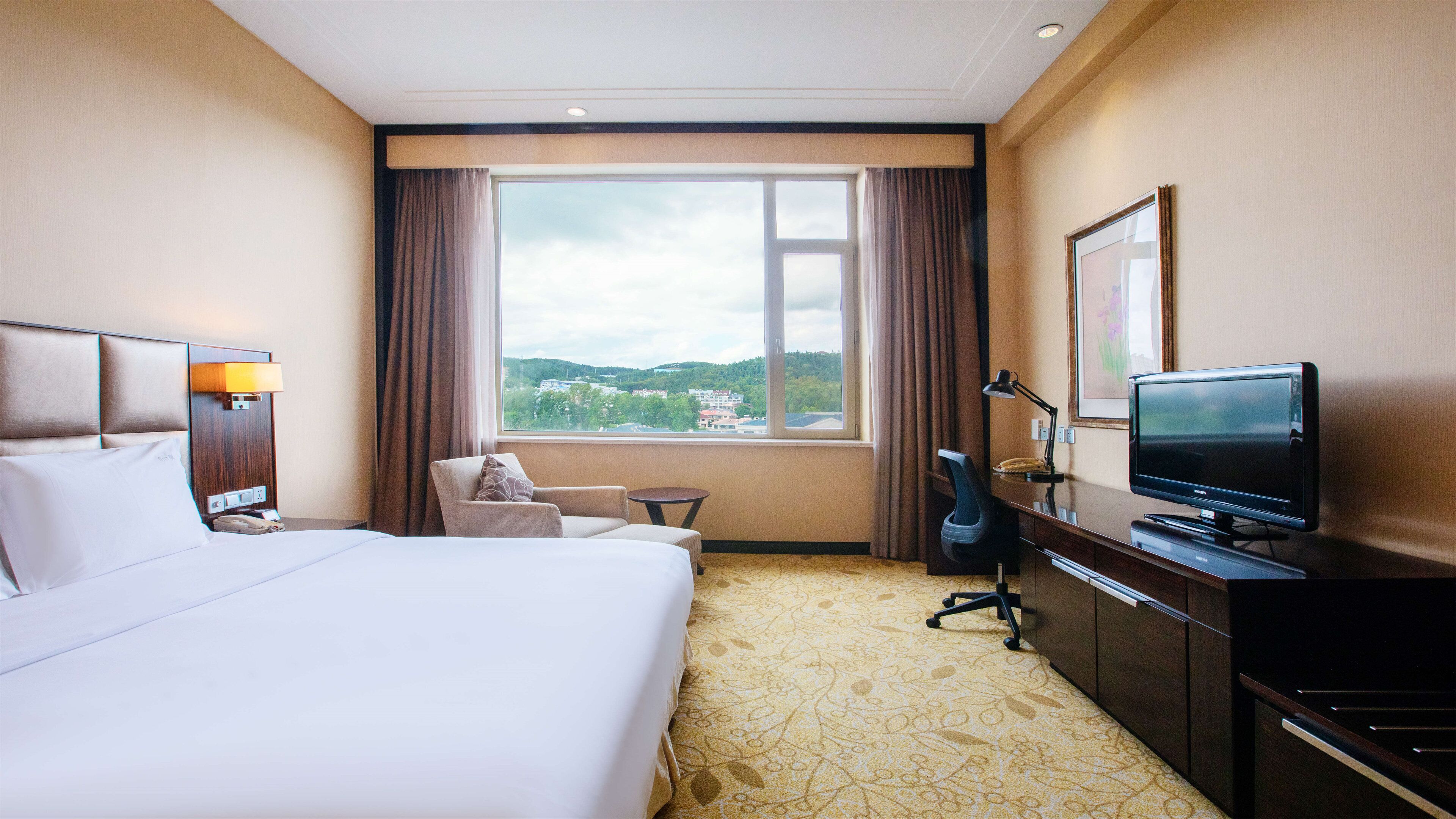 A hotel room with a bed, a desk with a television, a chair, a small table, and a window with a view of the city.