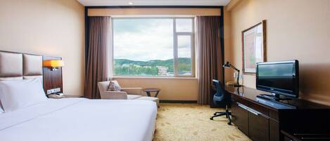 A hotel room with a bed, a desk with a television, a chair, a small table, and a window with a view of the city.