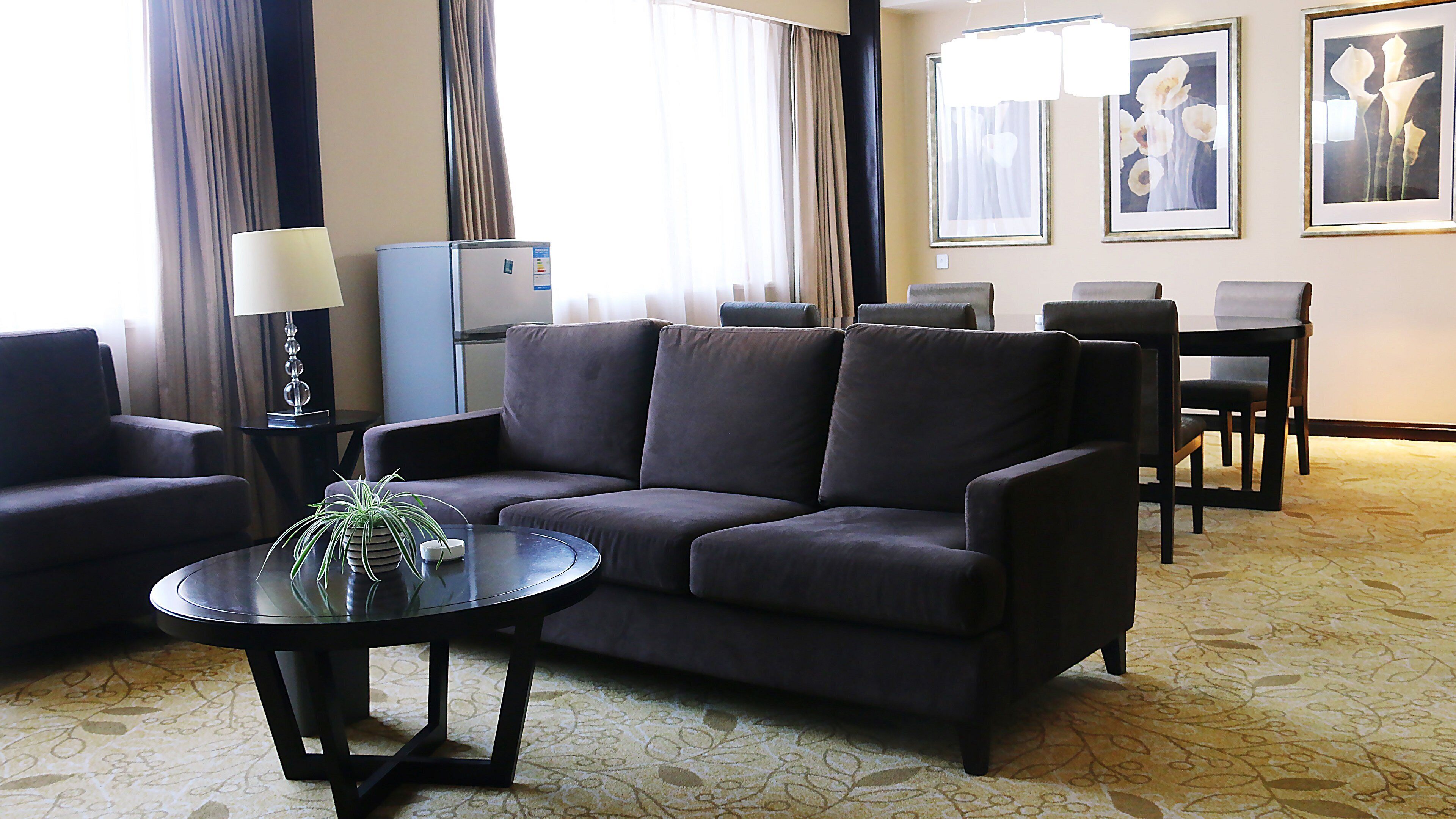 A hotel room with a dark-colored sofa, a small round table with a plant, a lamp, and a minibar. There are two framed pictures on the wall and a carpeted floor.