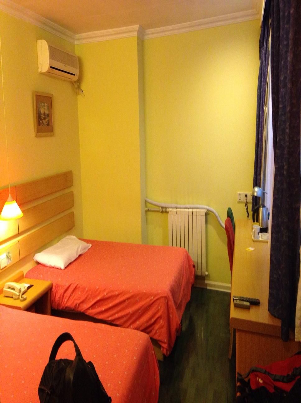Ein Hotelzimmer mit zwei Betten, einem Heizkörper, einem Nachttisch mit Lampe und einem Koffer auf dem Boden.