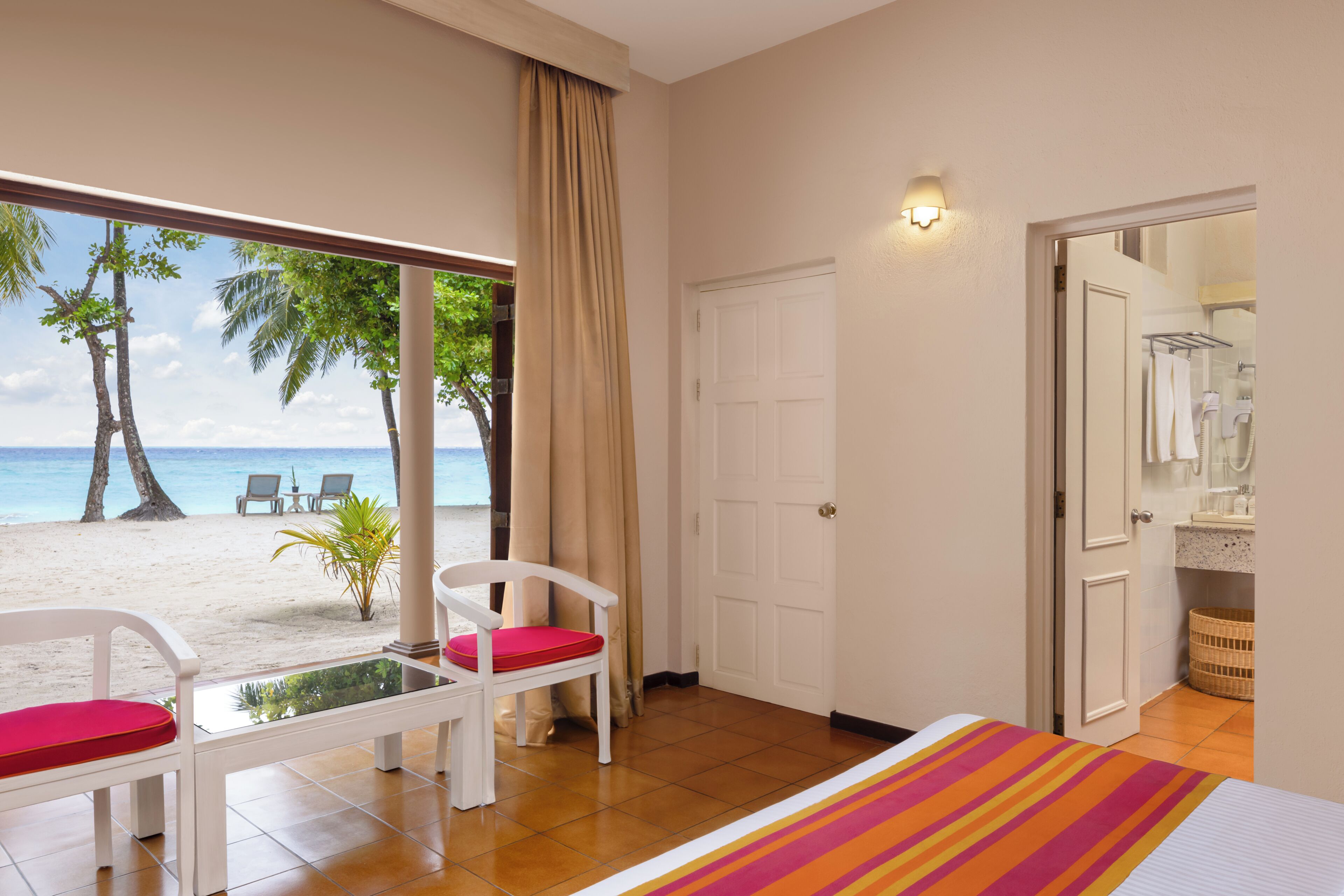 Une chambre d’hôtel avec vue sur la plage, un lit, une fenêtre avec des rideaux, une porte blanche et une salle de bain visible à travers une porte ouverte.