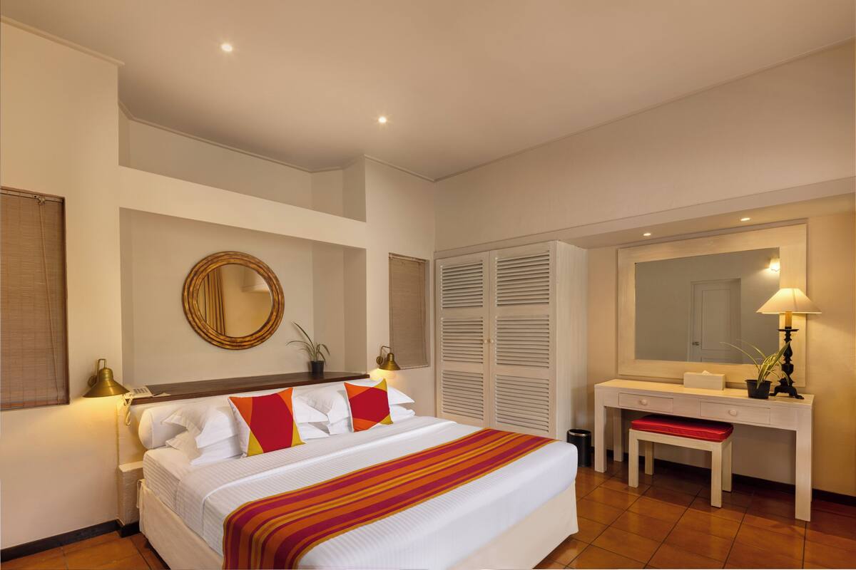 A hotel room with a bed, bedside table, lamp, mirror, and a desk with a chair.
