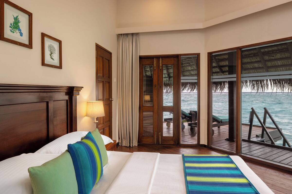 A hotel room with a bed, a view of the ocean, a wooden deck, and a thatched roof structure.