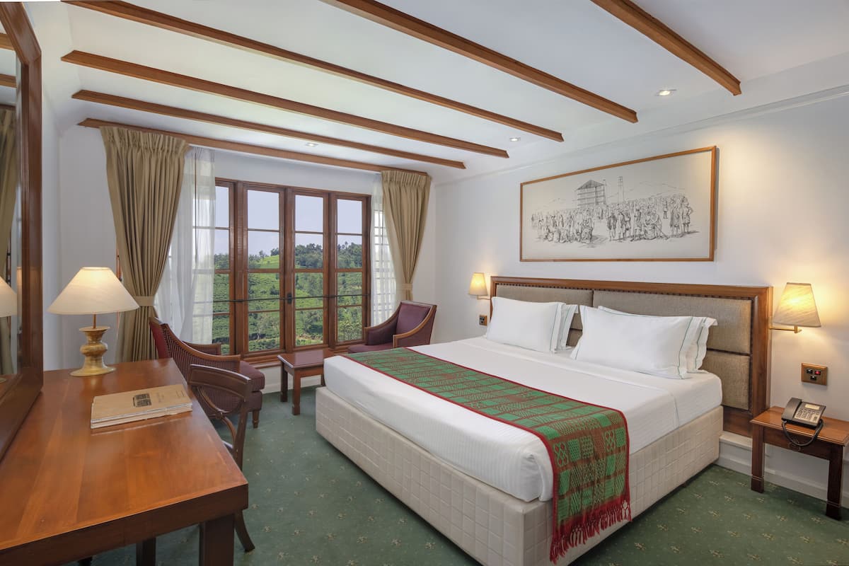 A hotel room with a large bed, a desk with a lamp, a chair, and a window with curtains.