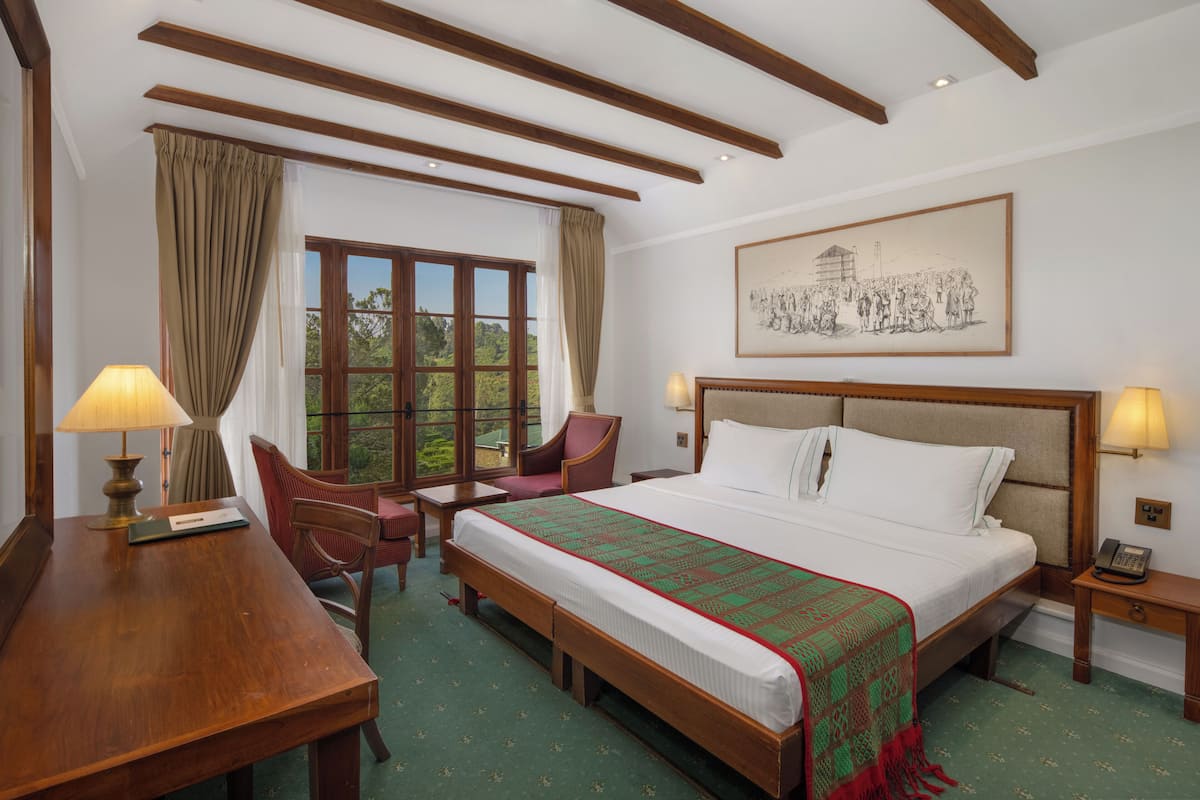 A hotel room with a large bed, wooden headboard, two bedside tables with lamps, a desk with a chair, and a window with curtains.