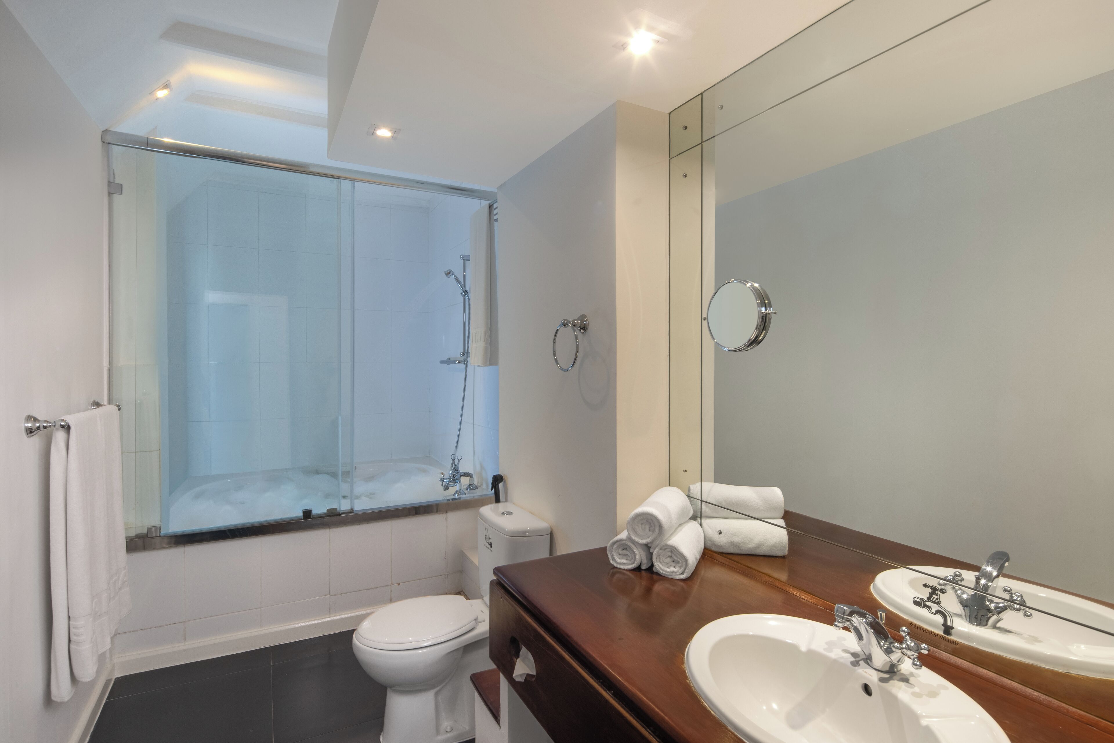 Un baño moderno con ducha de vidrio, inodoro blanco, tocador de madera con lavamanos y espejo, y toallas enrolladas ordenadamente.