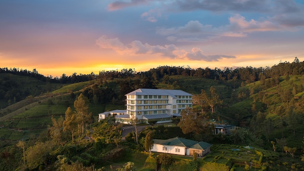 A large, multi-story hotel with a flat roof, surrounded by lush green hills and a sunset sky. - Heritance Tea Factory (Kandapola)