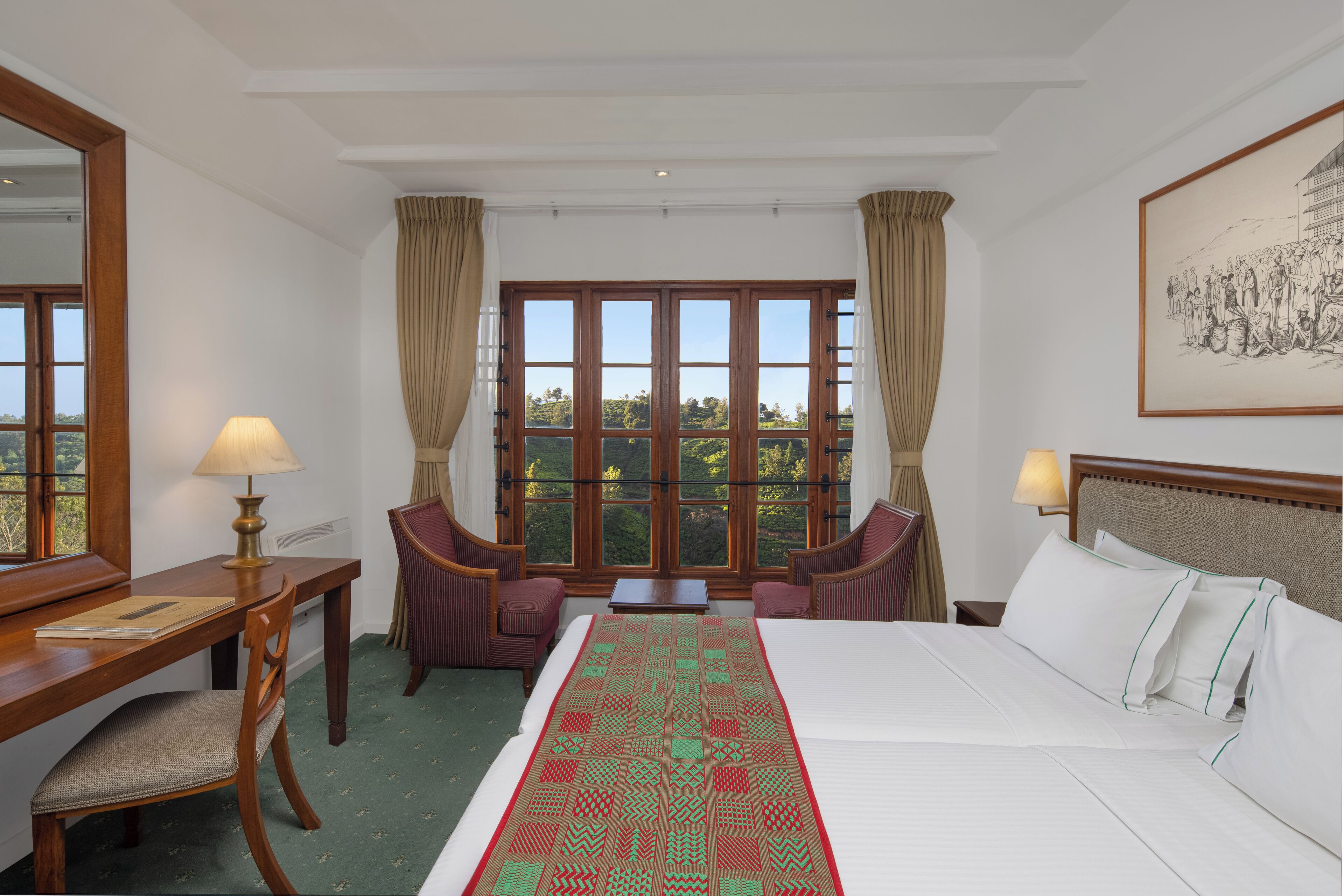 Een hotelkamer met een groot bed, een bureau met lamp, twee stoelen, een raam met gordijnen en een ingelijste foto aan de muur.