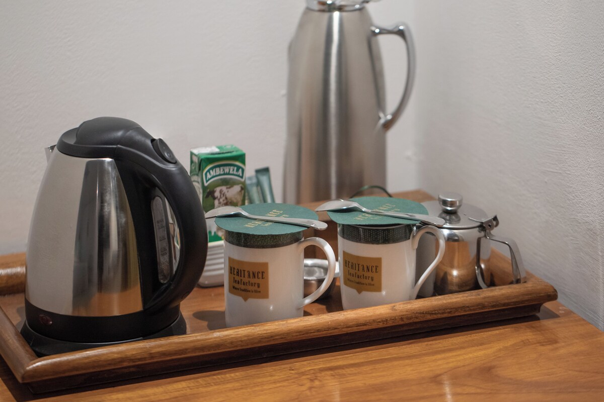 Un plateau avec une cafetière, deux tasses portant l’inscription « Heritage Tea Factory », une boîte de thé « Ambewela » et une théière en argent.