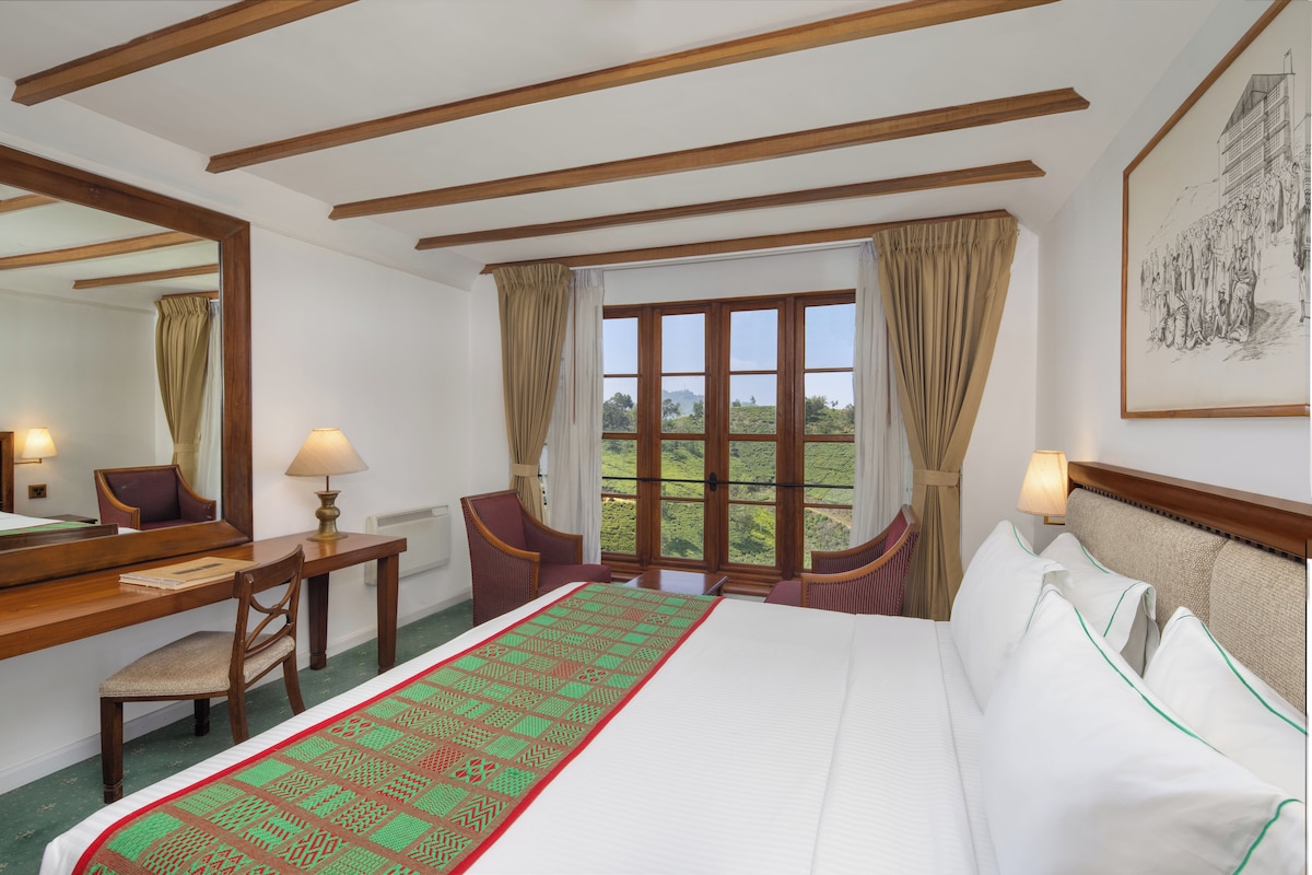 Quarto de hotel com uma cama grande, uma escrivaninha, duas cadeiras, uma janela com cortinas e um quadro na parede.