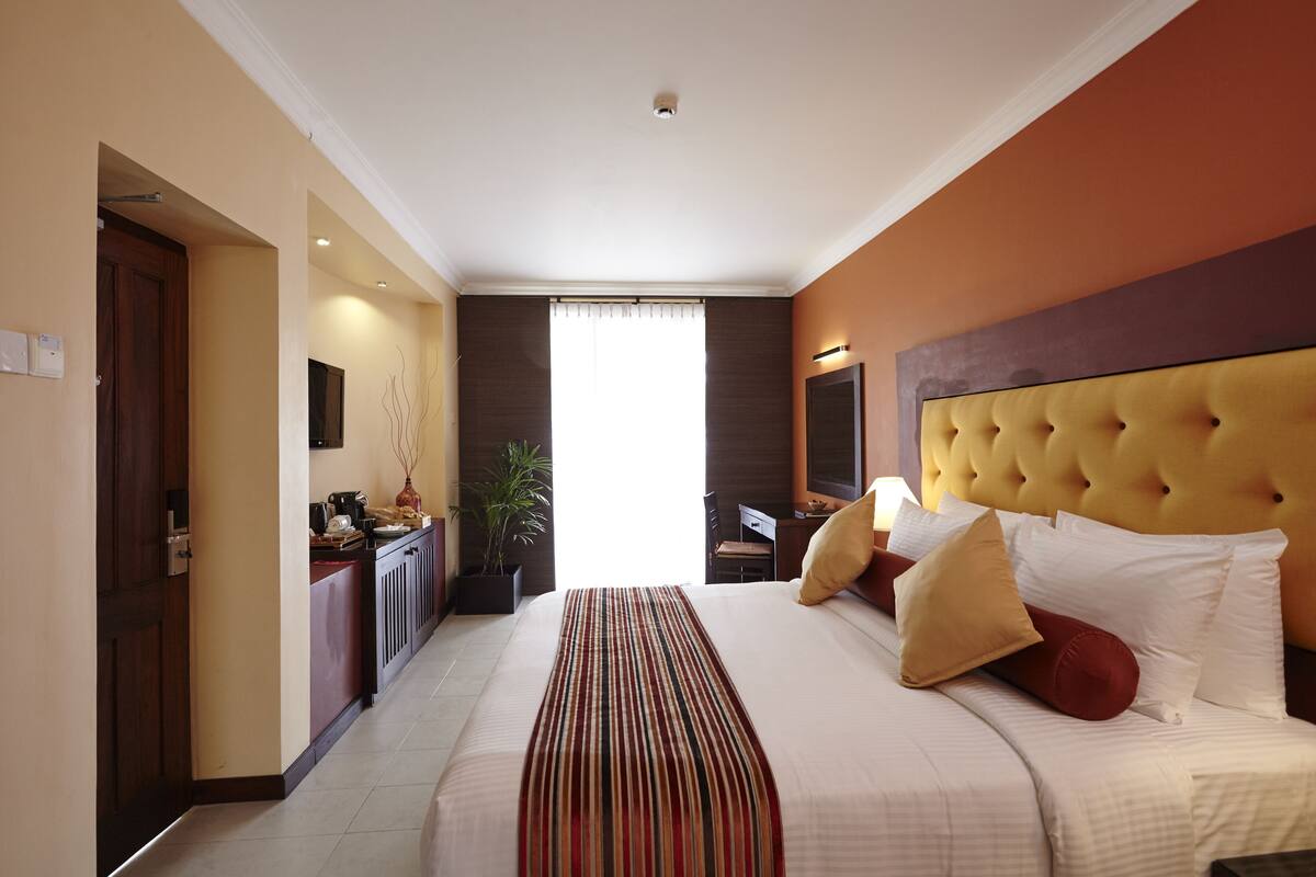 Quarto de hotel com uma cama grande, cabeceira, escrivaninha, cadeira, planta e janela com cortinas.