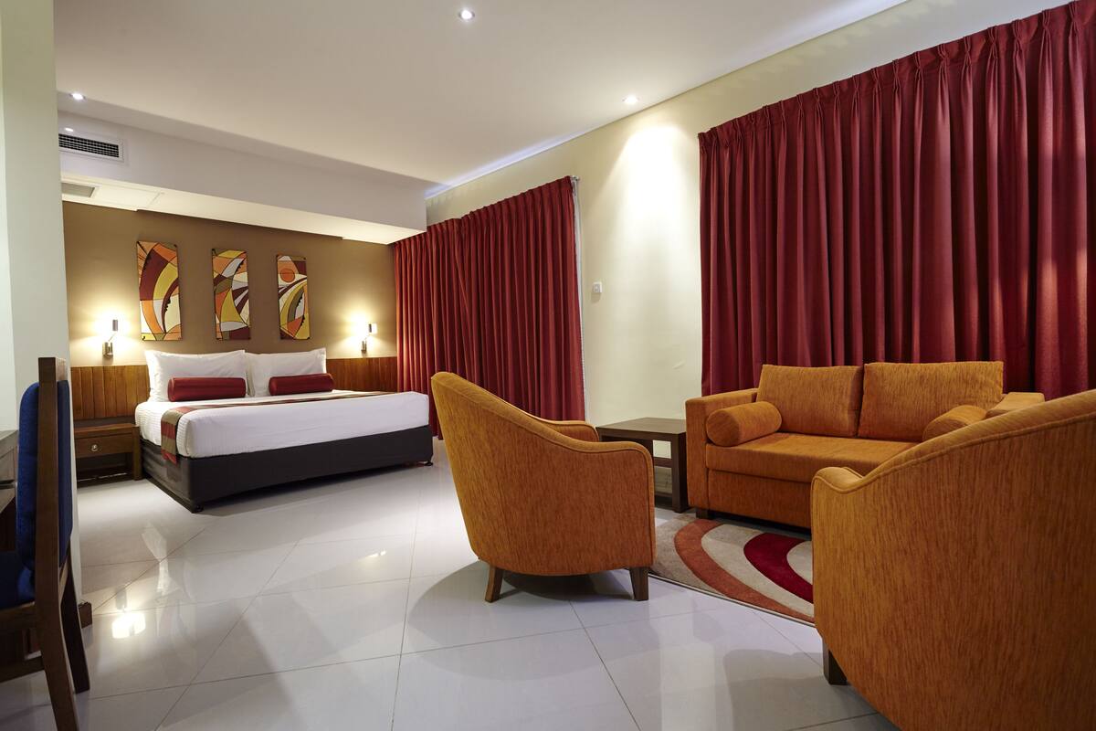 Quarto de hotel moderno com cama, duas poltronas, um sofá, uma mesinha e uma cortina vermelha.