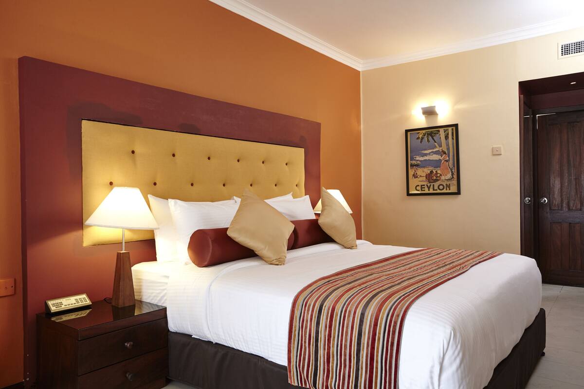 Quarto de hotel com uma cama grande, uma mesa de cabeceira com abajur e um quadro pendurado na parede.
