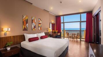 A hotel room with a large bed, a desk with a chair, a balcony with a view of the sea, and artwork on the wall.