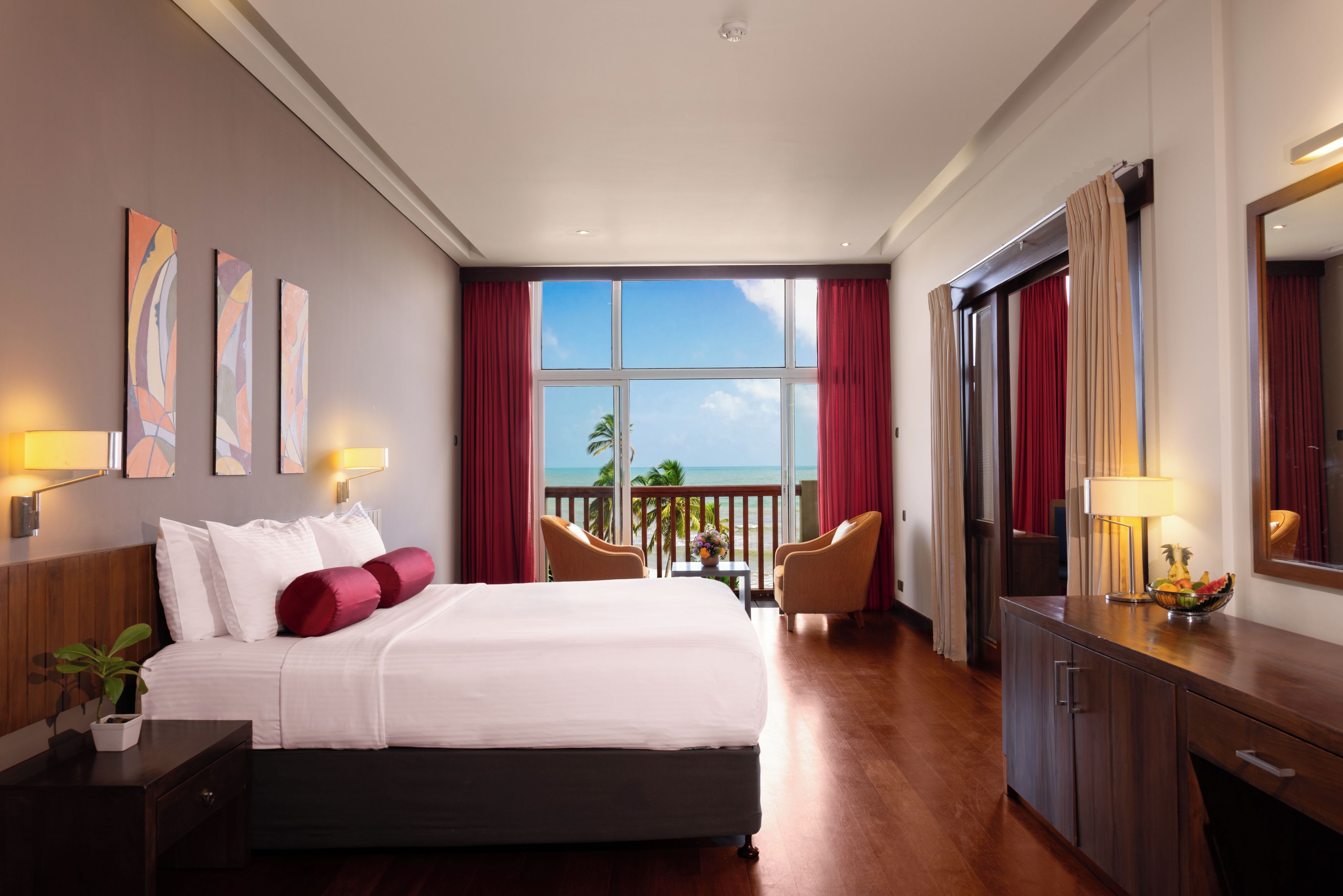 A hotel room with a bed, a desk, a chair, a large window with a view of the ocean, and artwork on the wall.