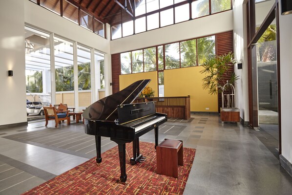 A grand piano in a spacious lobby with large windows, a wooden bench, and a potted plant. - Turyaa Kalutara (Wadduwa)