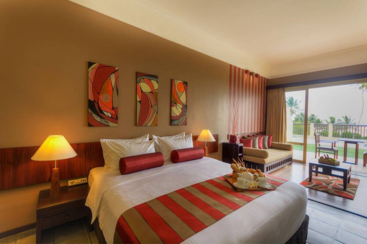 Quarto de hotel com uma cama grande, duas luminárias de cabeceira, um sofá e uma mesa de jantar. Há três quadros abstratos na parede e uma vista do exterior pela janela.