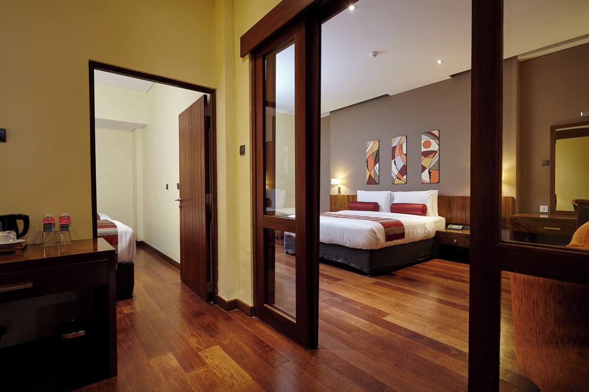 Quarto de hotel com piso de madeira, cama com roupa de cama vermelha e branca, uma escrivaninha com cadeira e um espelho.
