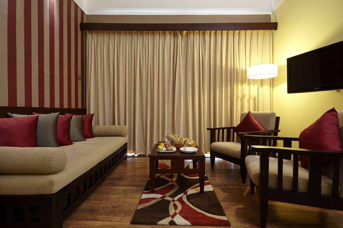 Quarto de hotel com sofá, poltronas, mesa de centro com frutas e televisão.