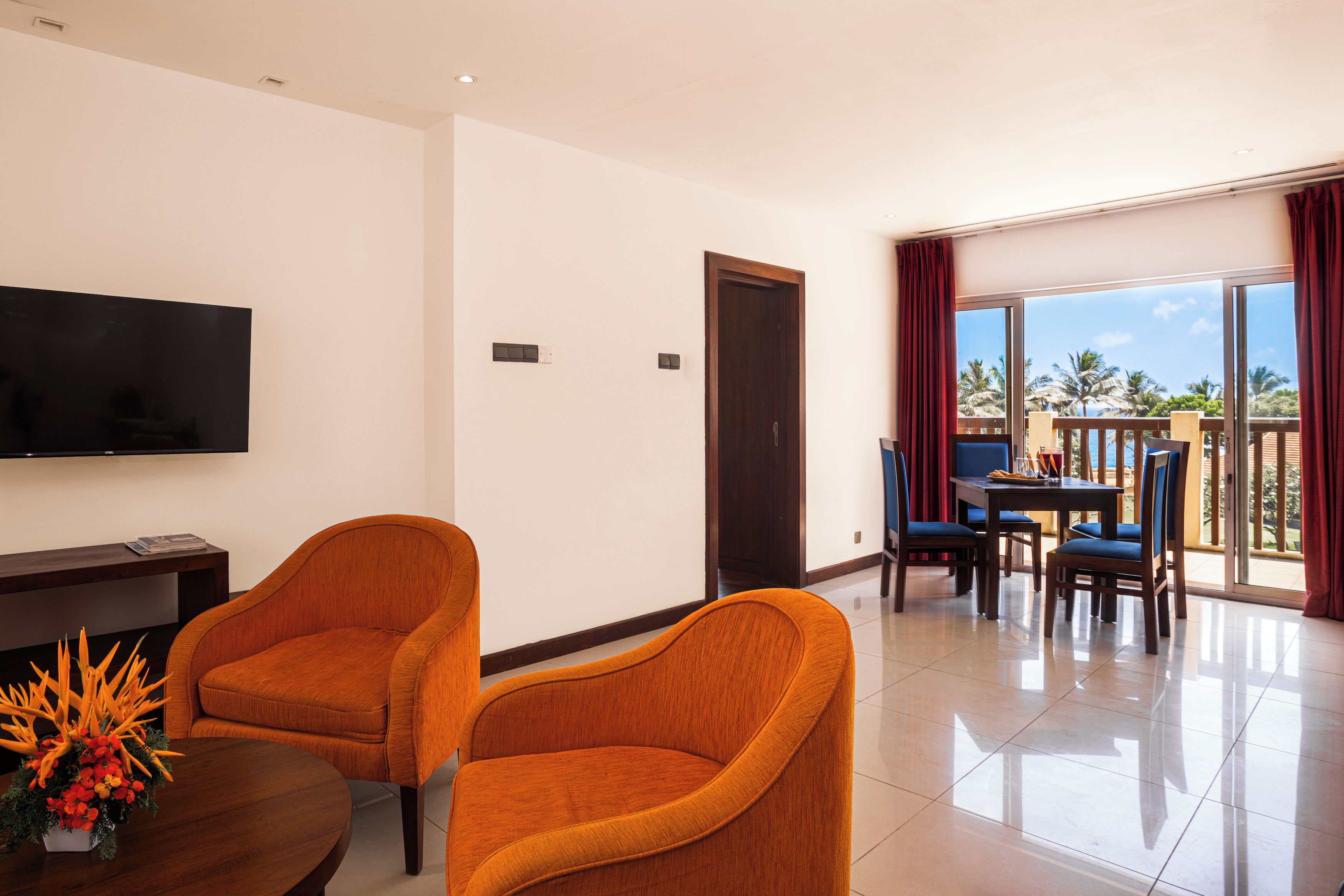 A modern living room with two orange armchairs, a flat-screen TV, and a dining area with blue chairs.