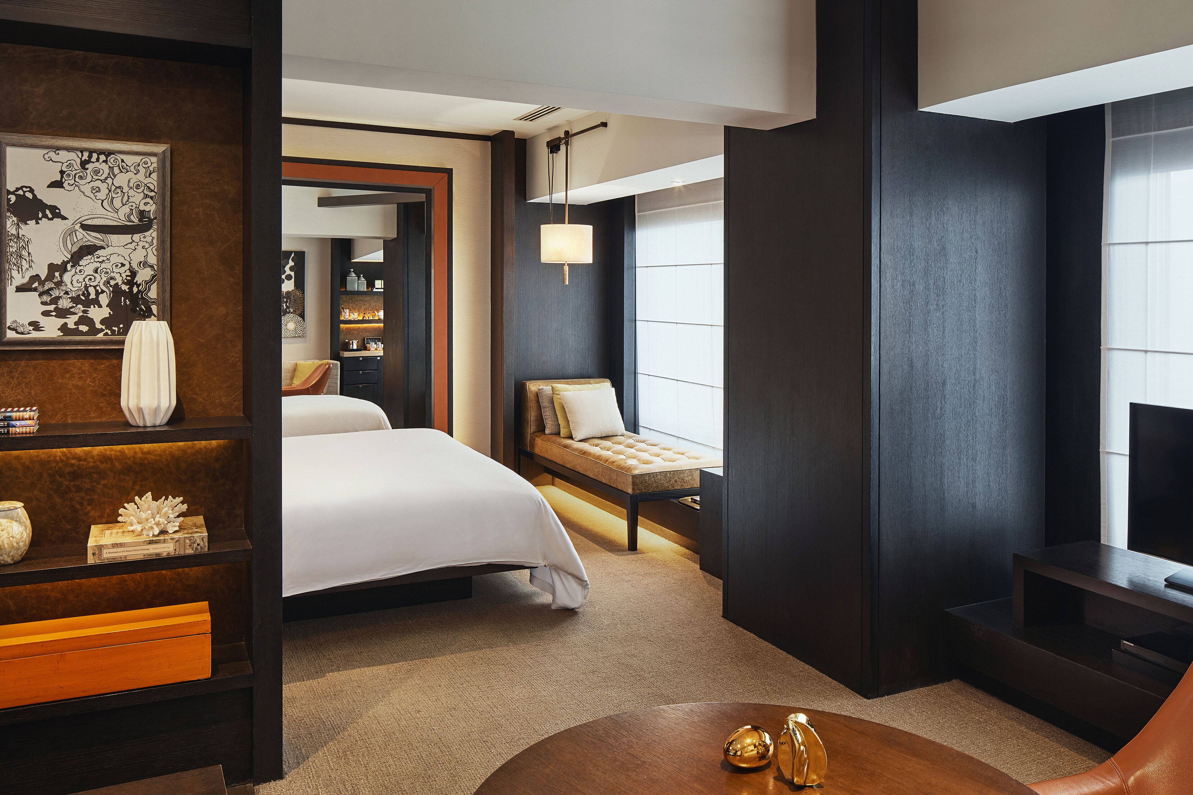 A modern hotel room with a large bed, a seating area with a sofa and armchair, a small table with decorative items, a bookshelf, and a large window providing natural light.