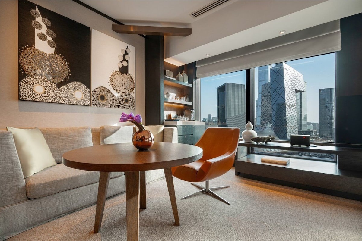 A modern living room with a round wooden table, a sofa, and a chair. There is a large window with a city view.