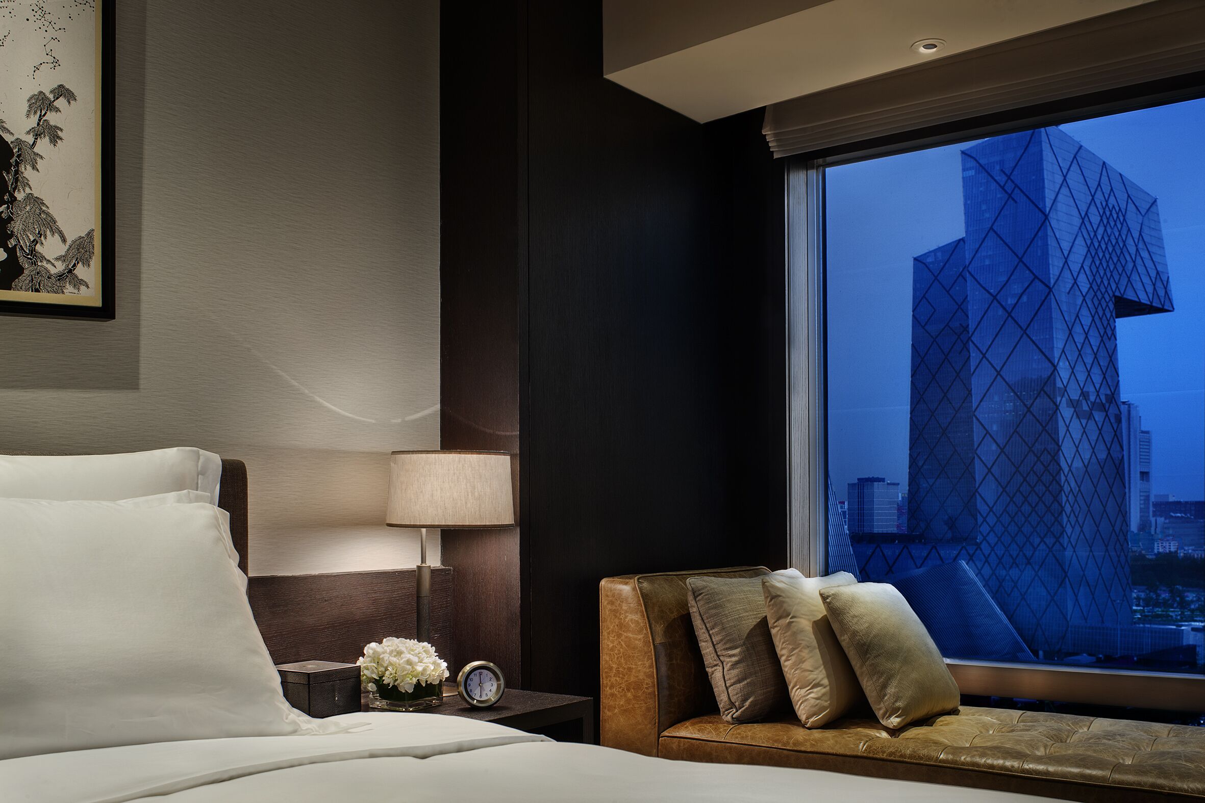Una habitación de hotel con una cama, una mesita de noche con lámpara, una pequeña mesa con flores y vistas a un edificio moderno a través de la ventana.