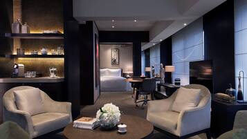 A modern hotel room with a bed, two armchairs, a coffee table with a vase, and a desk with a lamp.