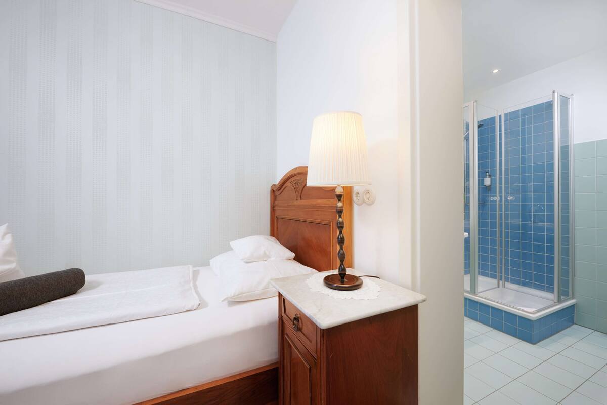 A hotel room with a bed, a nightstand with a lamp, a wooden headboard, a bathroom with a shower and blue tiles.