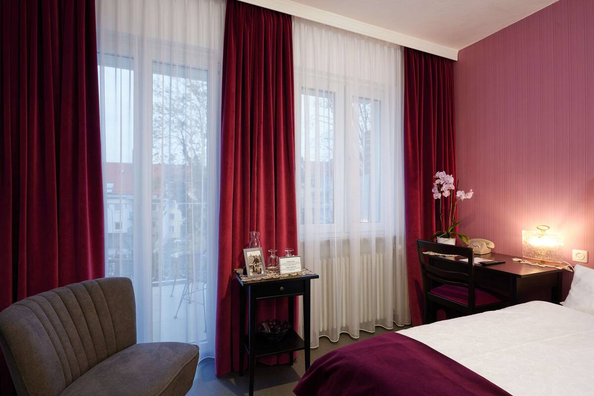 A hotel room with a bed, a desk with a telephone, a chair, a small table with a vase of flowers, and a window with red curtains.